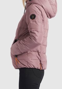 Vadderad jacka i mjuk rosa med huva, sidofickor och en svart logotypapplikation på ärmen. Har kontrasterande dragkedjor i orange.