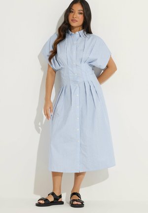 Kobieta w jasnoniebiesko-białej pasiastym sukience midi z krótkimi szerokimi rękawami i czarnych sandałach z klamrą, stojąca na jednolitym białym tle.