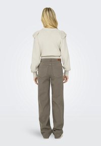Femme aux longs cheveux blonds, portant un pull beige avec des détails aux épaules et un pantalon gris ample, se tenant de dos sur un fond uni.