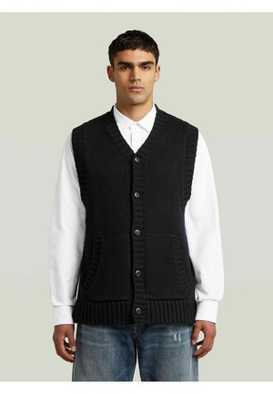 Giovane uomo che indossa un gilet nero lavorato a maglia abbottonato sopra una camicia bianca a maniche lunghe e jeans blu, in piedi davanti a uno sfondo chiaro e uniforme.