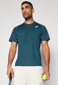Teal sportshirt met korte mouwen, knoopsluiting, textuurpatroon en grijze accenten. Model houdt een tennisbal en racket vast.