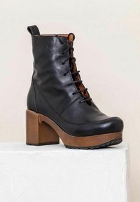Black leather ankle boot with a lace-up front, brown block heel, textured sole, and a smooth finish. Accented with a brown inner lining.