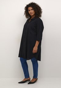 Chemise tunique noire à boutons sur le devant, à manches trois-quarts, en tissu texturé, coupe droite, associée à un jean skinny bleu et des talons escarpins noirs.