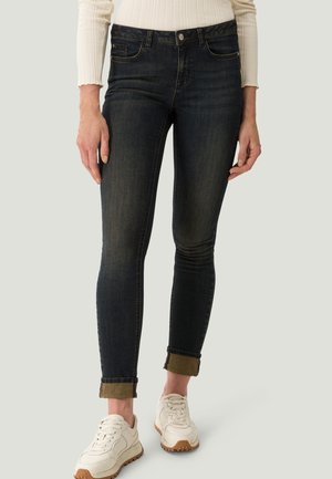 Persoon die zwarte strakke jeans met opgerolde pijpen draagt, een off-white geribd langarmshirt en witte sneakers met beige zolen, staand tegen een effen achtergrond.