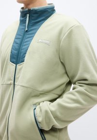 Ελαφρύ πράσινο μπουφάν φλις με ψηλό γιακά, διαθέτει μπροστινό φερμουάρ μπλε χρώματος και αντίθετες ενότητες. Περιλαμβάνει δύο πλαϊνές τσέπες και λογότυπο.