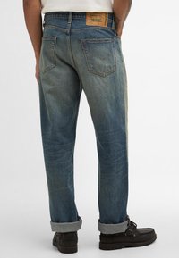 Jeans de denim azuis com um acabamento desbotado, apresentando um rótulo de couro, punhos enrolados e design de cinco bolsos. Acentos costurados e marcas de desgaste visíveis.