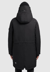 Man in een lange, zwarte, gevoerde winterjas met capuchon en een zwarte beanie, staand met zijn rug naar de camera tegen een effen grijze achtergrond.