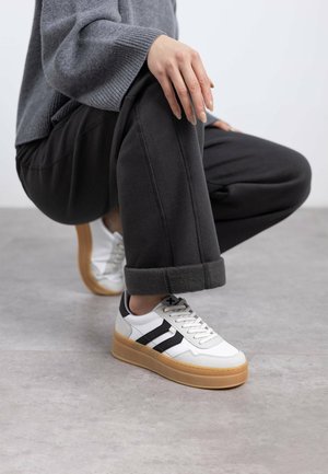 Zapatillas de cuero blancas con rayas negras, suela de goma elevada, puntera redonda y costuras en contraste. Llevadas con un suéter gris y pantalones negros.