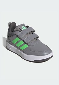 Zapatilla deportiva gris con detalles verdes, correas de velcro, collar acolchado, parte superior texturizada y suela de goma. Diseñada para brindar comodidad y soporte.