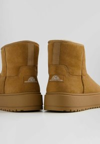 Bruna mockasiner med tjocka gummisulor och foder av päls, visande bakvy med "Nice & Easy Footwear" logotyp.