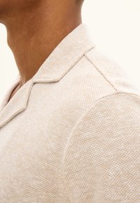 Närbild av en persons nacke och axel som bär en beigemelerad stickad tröja med en vid krage mot en neutral bakgrund.