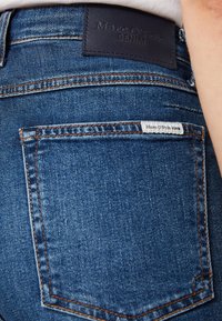 Jeans de mezclilla azul oscuro presentan una etiqueta de cuero en la cinturilla que dice "Marc O'Polo DENIM" y una pequeña etiqueta blanca en el bolsillo trasero.