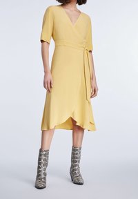 Gul omlottklänning med korta ärmar och ett knyt i midjan. Längden går nedanför knäet, med en svävande fåll. Buren med silvriga boots i ormtryck.