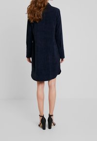 Mörkblått mockaklänning med lång ärm, rundad nederkant och sidoklyftor. Matchad med svarta högklackade sandaler.