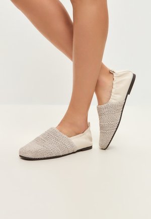Frauenbeine mit cremefarbenen Slip-On-Flats mit gewebter Textur vorne und glattem Stoff an der Ferse, stehend auf einem hellen Boden.