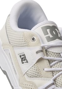 Vit sportskor med mesh och mocka material. Har grå detaljer, en vadderad tunga och texturerade inslag. DC-logotypen på sidan.