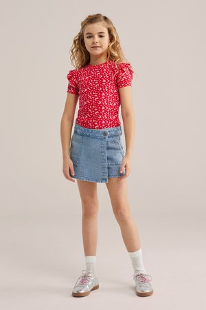 Meisje staand, gekleed in een rood patroon kortermig shirt, een denim wikkelrok, witte sokken en zilveren veterchoenen, tegen een effen achtergrond.