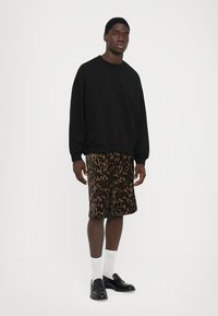 Jeune homme debout dans un sweat-shirt noir, un short imprimé léopard marron, des chaussettes blanches et des mocassins noirs devant un fond blanc uni.