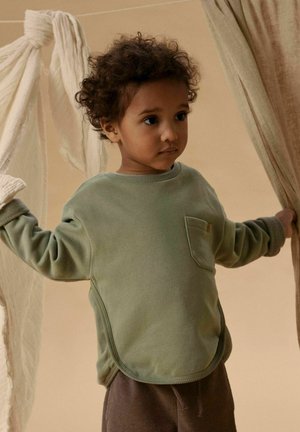 Nuori lapsi kiharat hiukset, pukeutunut vihreään collegepaitaan ja ruskeisiin housuihin, pitää vaaleanbeigeä kangasta molemmissa käsissään sisätiloissa.