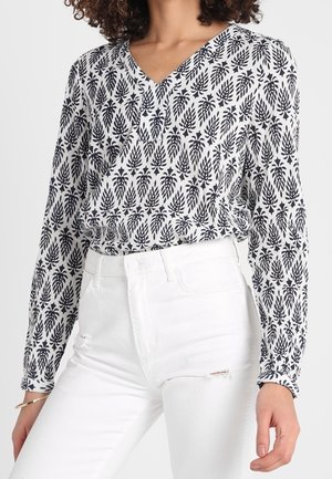Femme portant un chemisier à manches longues à motif feuilles noir et blanc rentré dans un jean blanc taille haute légèrement usé.