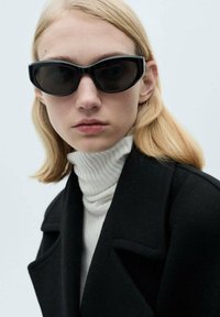 Črne sončne očali z angularnim dizajnom, temna leča, jih nosi oseba s straight blond lasmi, oblečena v črn plašč in belo turtleneck.