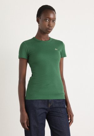T-shirt verde a coste con maniche corte, caratterizzato da un piccolo logo ricamato sul lato sinistro. Il tessuto è liscio e aderente, abbinato a jeans scuri.