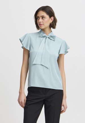 Blusa azzurra chiara con maniche corte a volant e colletto annodato. Tessuto morbido e vestibilità comoda. Abbinata a pantaloni neri.
