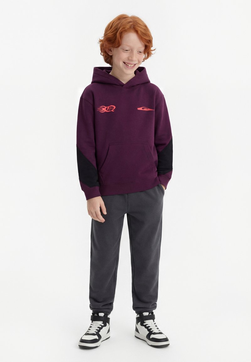 Punapäine poiss, kes kannab lillat kapuutsiga pusa punaste leegimustritega, tumedat halli pükse ja must-valgeid kõrgeid tossusid, naeratab ja vaatab allapoole.