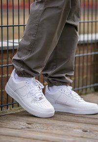 Bílé tenisky Nike Air Force 1 s perforovanou špičkou, plochými tkaničkami a strukturovaným koženým povrchem, spárovány s volnými béžovými kalhotami.