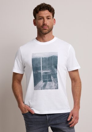 Homme aux cheveux courts et bouclés portant un t-shirt blanc à motif graphique bleu abstrait, debout avec une main dans la poche sur un fond uni.