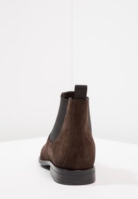 Botín Chelsea de ante marrón con paneles laterales elásticos, una lengüeta y una suela plana de goma negra. El diseño es elegante y con un mínimo de detalles.