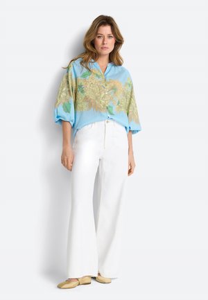 Frau trägt ein blaues, blumiges Hemd mit Knöpfen und Puffärmeln, weiße Hosen mit weitem Bein und beige Ballerinas, steht vor einem weißen Hintergrund.