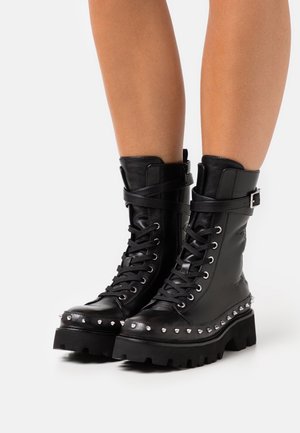 Musta nahkainen taistelukenkä, jossa on niittikoristeltu reunus, nauhallinen etuosa, solkisangat ja paksut nastapohjat, käytetty paljain jaloin.