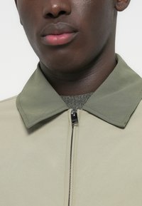 Κοντινό πλάνο ενός ατόμου που φοράει μια ανοιχτό μπεζ ζακέτα με πράσινο γιακά και ένα μερικώς ορατό γκρι πλεκτό ρούχο από κάτω.