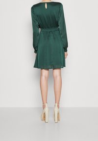 Grön satin klänning med långa ärmar, dragsko i midjan och nyckelhålsdetalj bak. Tillsammans med beige högklackade skor.