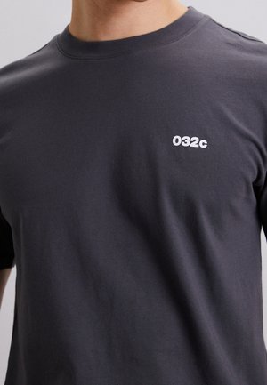 Czarnoszary bawełniany t-shirt z klasycznym okrągłym dekoltem i krótkimi rękawami. Posiada małe białe logo "032c" na górnej lewej części klatki piersiowej.