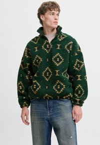 Zielony polarowy sweter z zamkiem przy kołnierzu, z geometrycznym brązowym i czarnym wzorem. Luźny krój z elastycznymi mankietami, miękka tekstura.