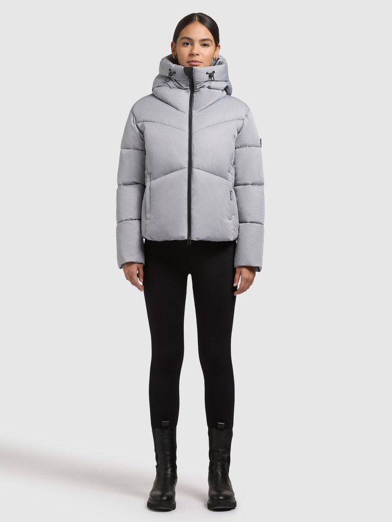 Sivá prešívaná bunda s vysokým golierom a kapucňou, vybavená predným zipsom a švami v tvare chevronu. Nosená na čiernych legínach a čižmách.