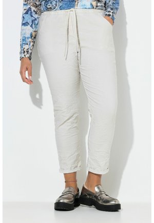 Persona che indossa pantaloni bianchi con coulisse e orli risvoltati, mocassini metallici e una maglia a maniche lunghe blu con motivo, in piedi contro una parete bianca.