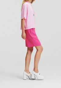 Žena v růžovém oversized tričku, jasně růžové sukni ke kolenům a bílých platformových botách na silném podpatku, stojící před jednobarevným pozadím.