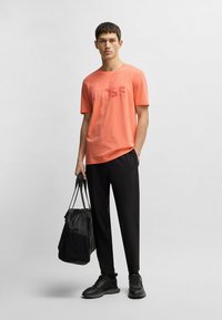 Koralfarvet bomulds-T-shirt med 'BOSS'-logo, parret med sorte bukser og sorte sneakers. Modellen holder en sort taske.
