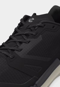 Κοντινό πλάνο μαύρου αθλητικού παπουτσιού με υφασμάτινη υφή, μαύρα κορδόνια και γκρι άκρη σόλας με μικρό λογότυπο στη γλώσσα του παπουτσιού.