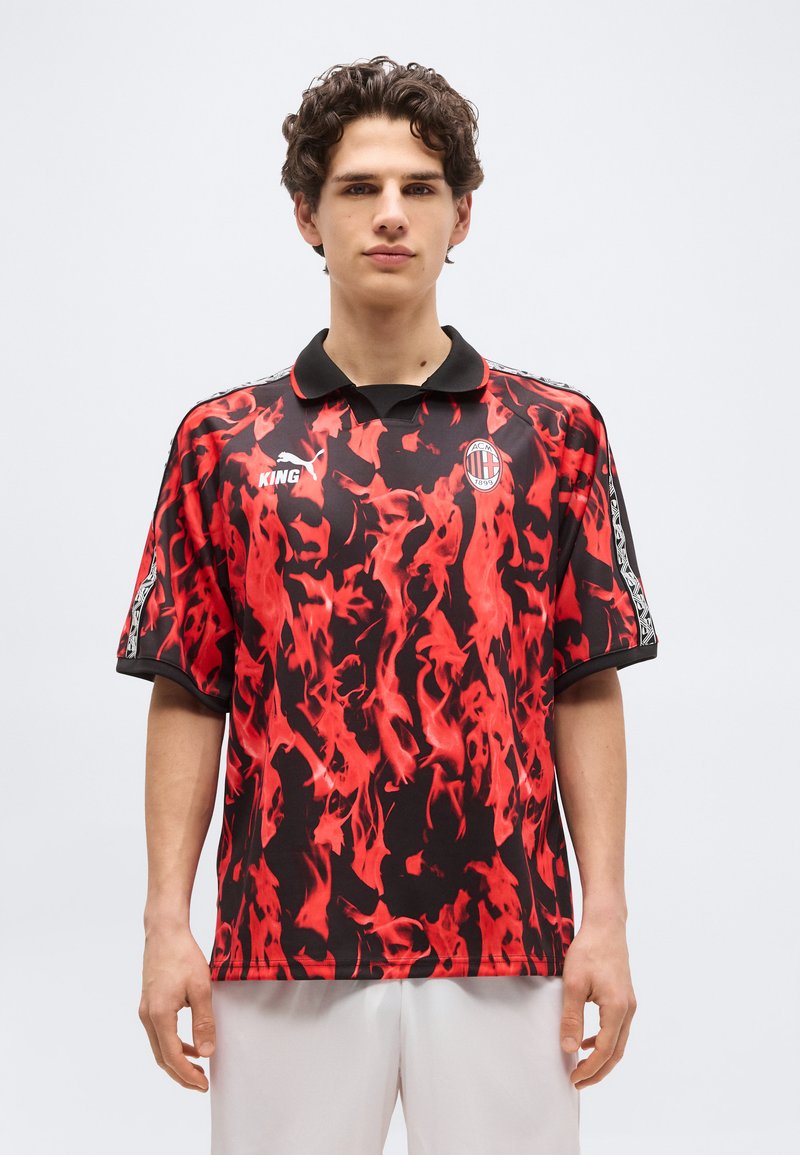 Νέος άνδρας με φανέλα ποδοσφαίρου της AC Milan με κόκκινο και μαύρο σχέδιο, μαύρο γιακά και λευκά σορτς, στέκεται μπροστά σε απλό φόντο.