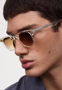 Ochelari de soare clar, supradimensionați, cu lentile degradate de culoare maro, rame transparente și detalii argintii pe brațe; modelul poartă o cămașă cu guler.