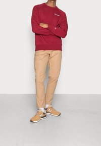 Felpa rossa con logo, pantaloni beige e sneakers marroni con dettagli bianchi. Tessuto liscio, vestibilità casual e polsini a coste.
