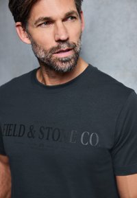 Tmavě šedé tričko vyrobené z bavlny, s velkým tištěným logem "FIELD & STONE CO." v různých odstínech šedé na přední straně.