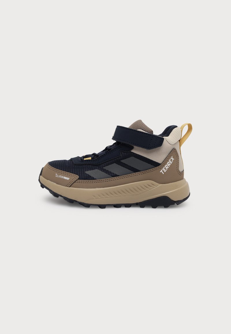 Παιδικό πεζοπορικό παπούτσι Adidas Terrex σε μαύρο και μπεζ με ρυθμιζόμενη λουρίδα, παχιά σόλα και κίτρινη ταινία έλξης στη φτέρνα.