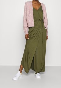 Maxi dress in olive green with draped design and side tie detail, paired with a chunky pink cardigan and white sneakers.