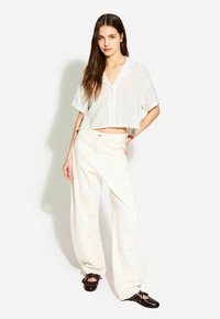 Camicia bianca a maniche corte con bottoni, abbinata a pantaloni bianchi a gamba larga e zoccoli marroni scuri con dettagli decorativi in metallo.