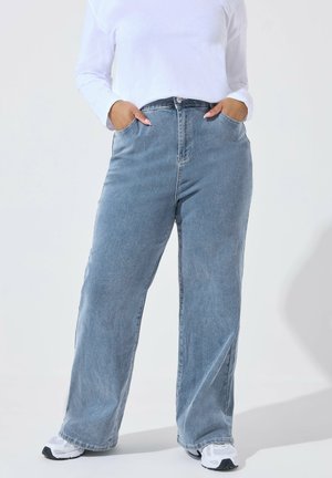 Person iført løstsiddende lyseblå jeans og hvide sneakers, hænder i lommerne, stående mod en ensartet lys baggrund.